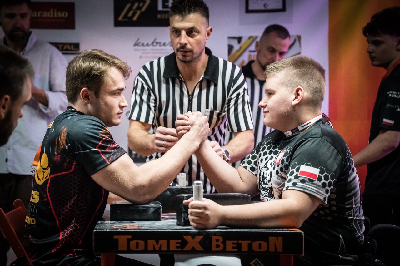 Na zdjęciu dwie osoby w trakcie rywalizacji w armwrestlingu oraz sędzia zawod&oacute;w